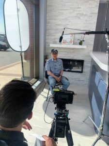 Person seated for an on-camera interview near the front window and fireplace at Avatar Dental, with lighting, microphone, and video equipment set up